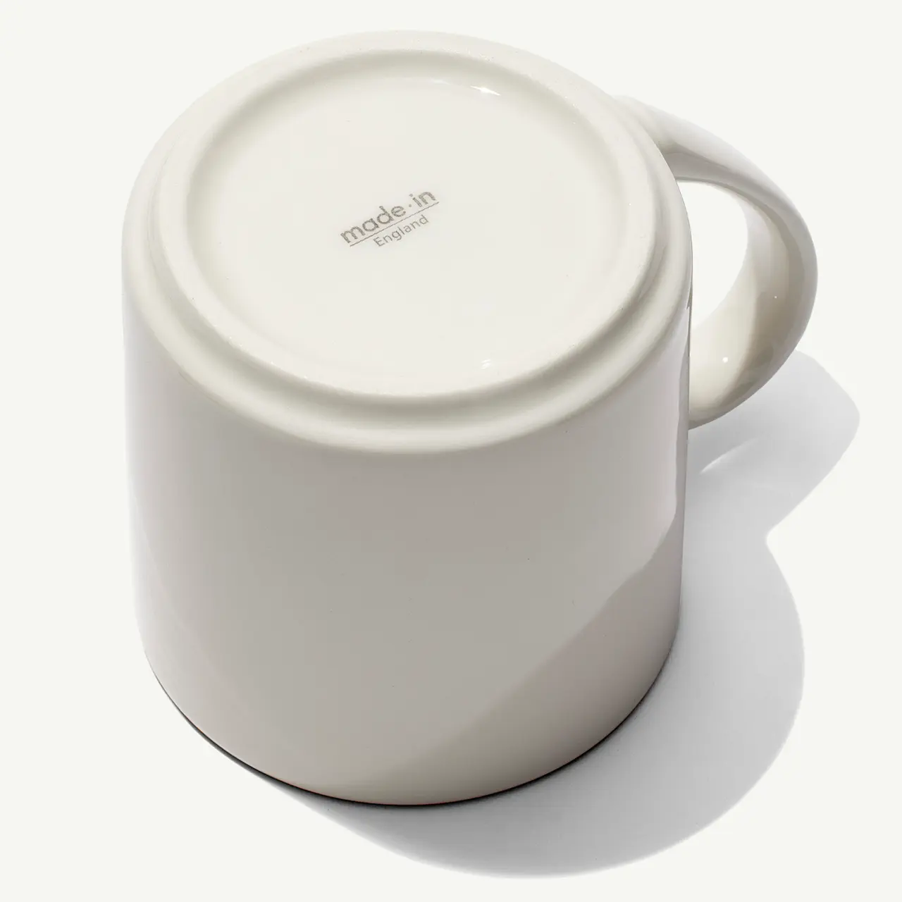 A white ceramic mug is positioned upside down on a white surface, with its handle to the left and text on the bottom indicating its brand or manufacturing origin.