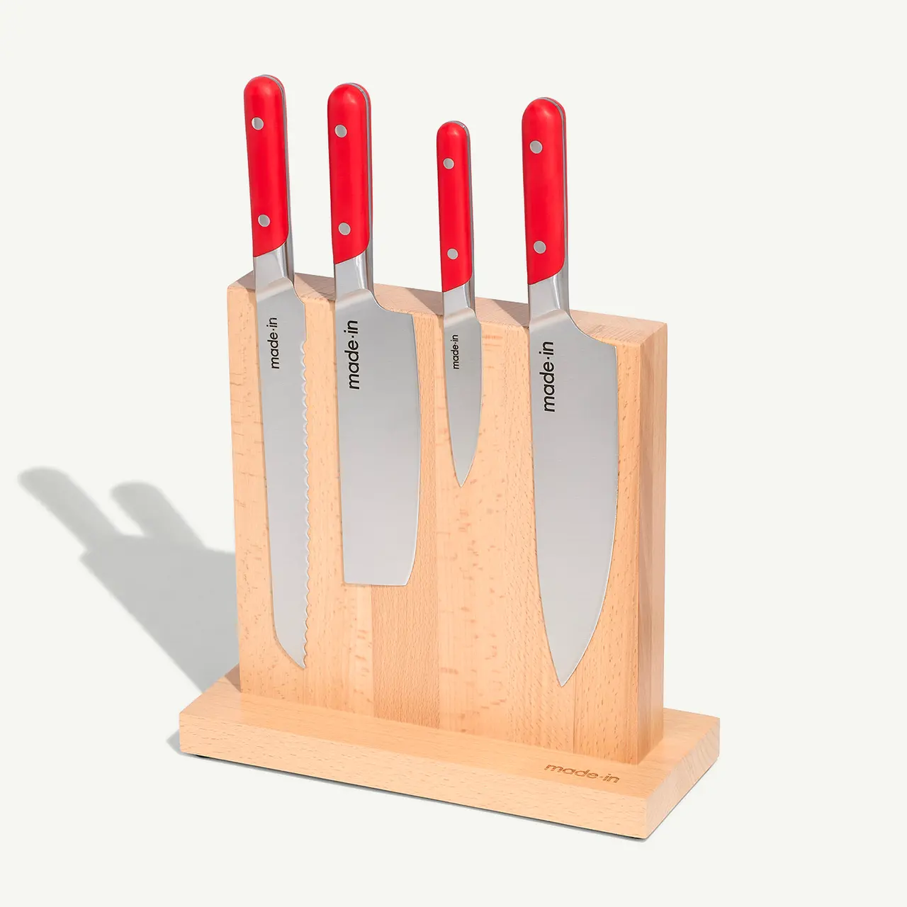 A set of four kitchen knives with red handles is neatly arranged in a light wooden knife block casting a shadow on the surface behind it.