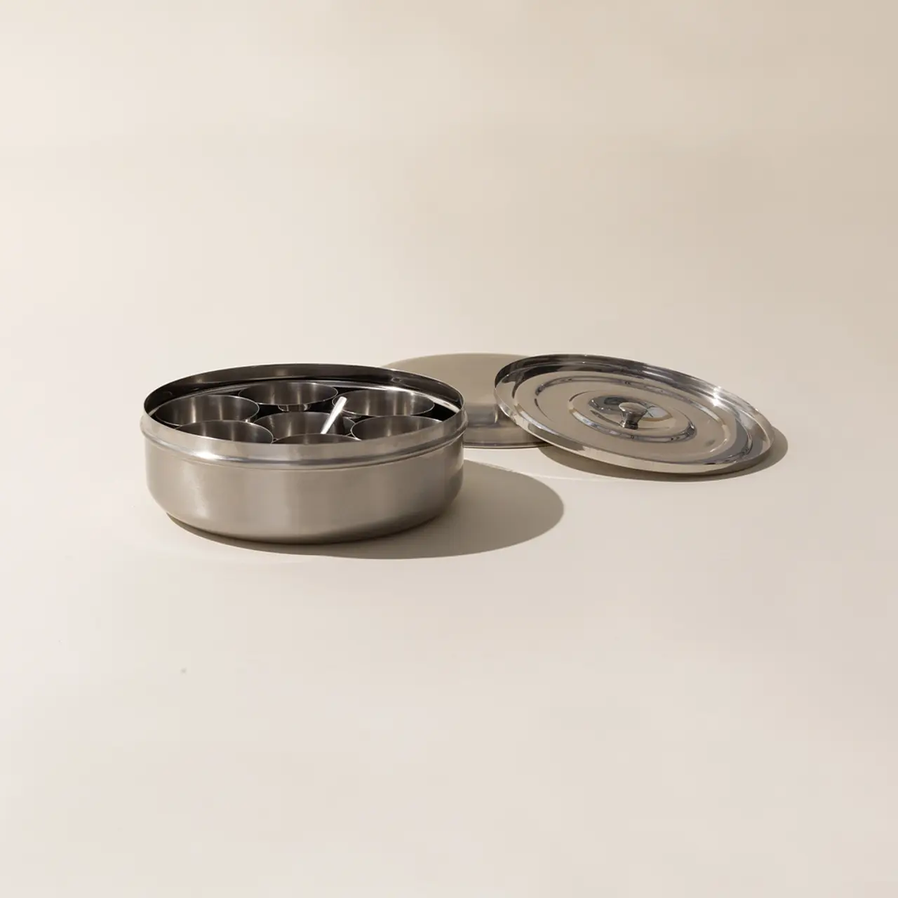 A stainless steel ashtray with multiple cigarette rests is shown beside its detached lid on a pale surface.