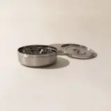 A stainless steel ashtray with multiple cigarette rests is shown beside its detached lid on a pale surface.