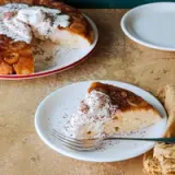 A slice of banana cake topped with whipped cream and cocoa powder alongside a whole cake on a red-rimmed plate.