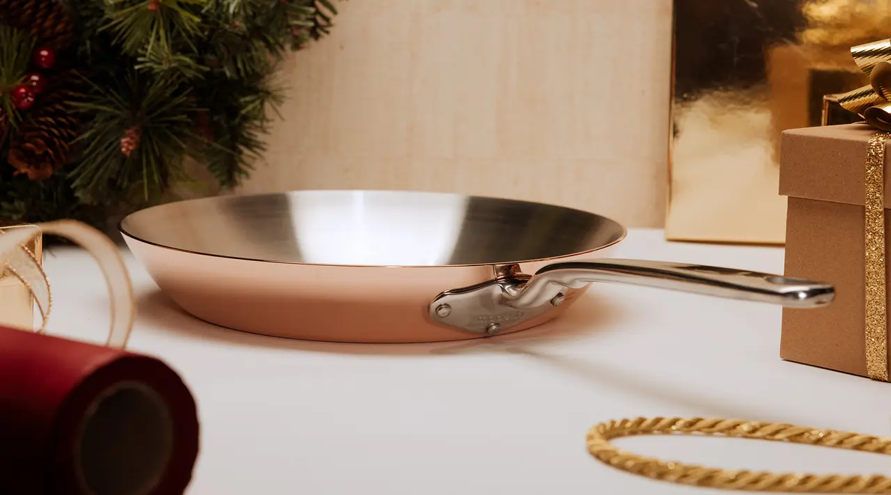 A stainless steel frying pan with a copper exterior is displayed on a table amidst holiday decorations and wrapped gifts.