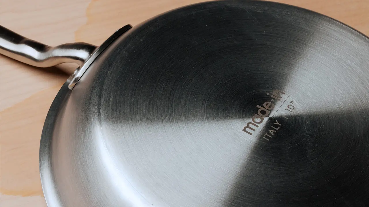 A stainless steel frying pan with a riveted handle is resting on a wooden surface.