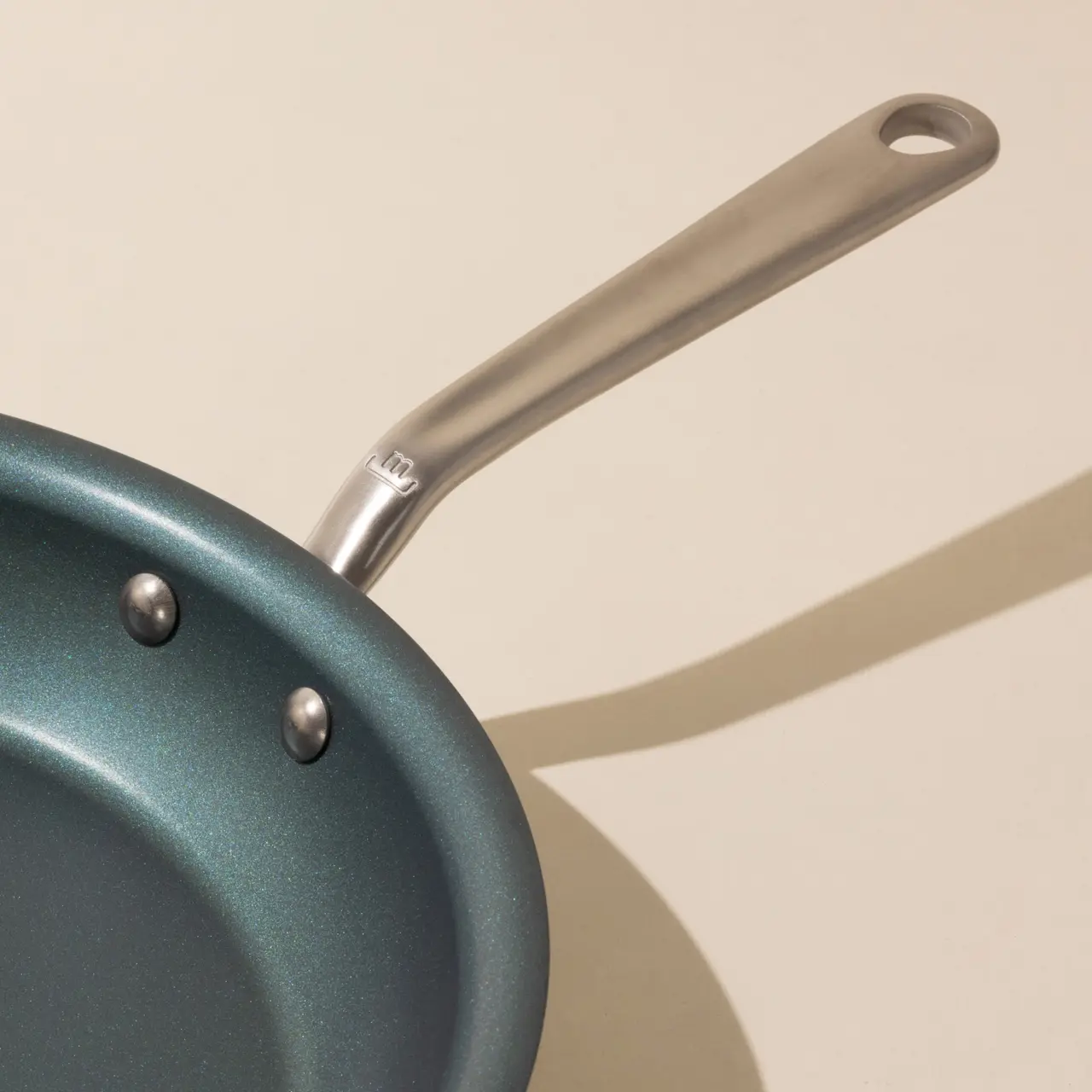 A blue non-stick frying pan with a stainless steel handle casts a shadow on a light surface.