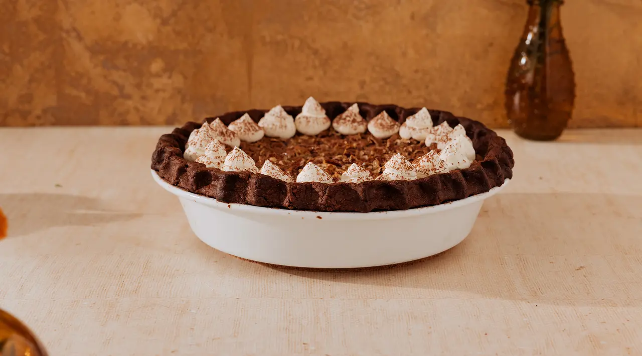 A chocolate pie topped with whipped cream dollops and a sprinkle of cocoa powder sits on a countertop with a rustic backdrop.