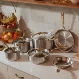 A set of stainless steel pots and pans with gold-colored handles is neatly arranged on a kitchen counter by a tiled backsplash, with a fruit basket nearby.