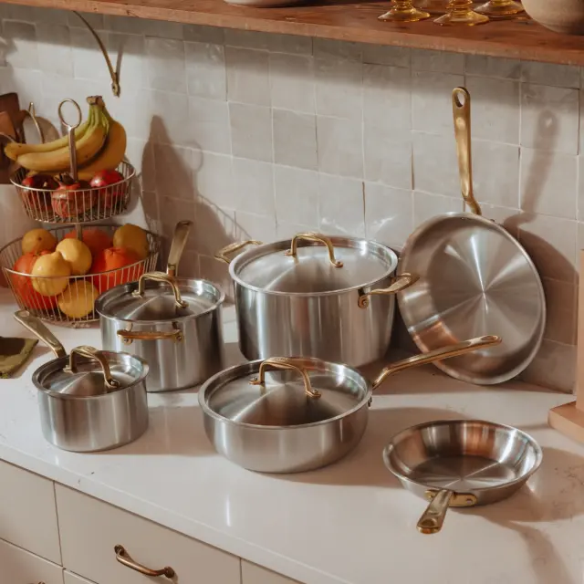 A set of stainless steel pots and pans with gold-colored handles is neatly arranged on a kitchen counter by a tiled backsplash, with a fruit basket nearby.