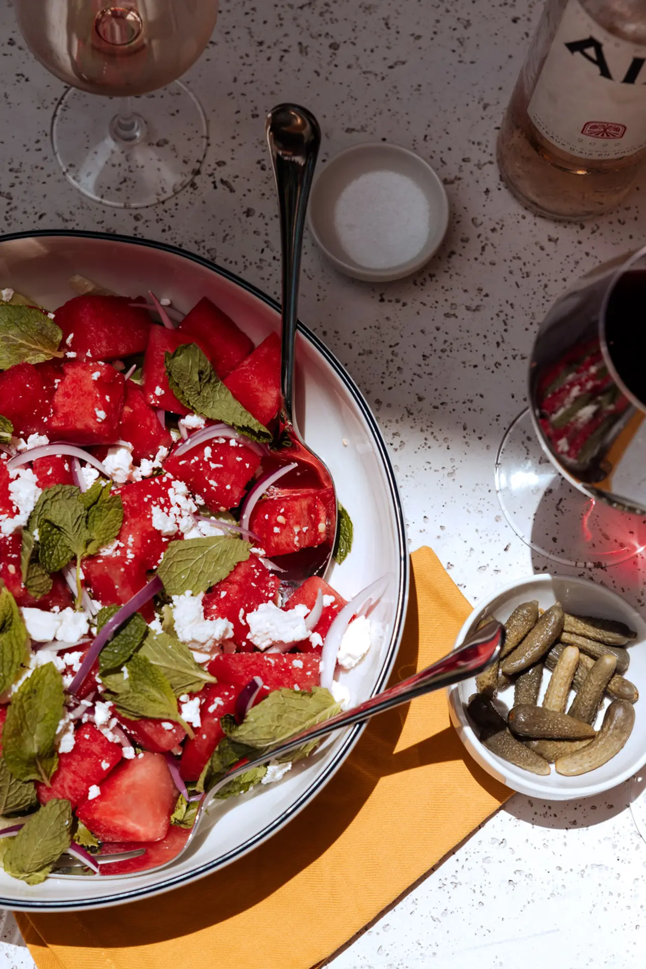 A bowl of watermelon salad sprinkled with feta cheese and mint leaves is accompanied by a small dish of pickles and a saltshaker, all set on a table with a glass of wine.