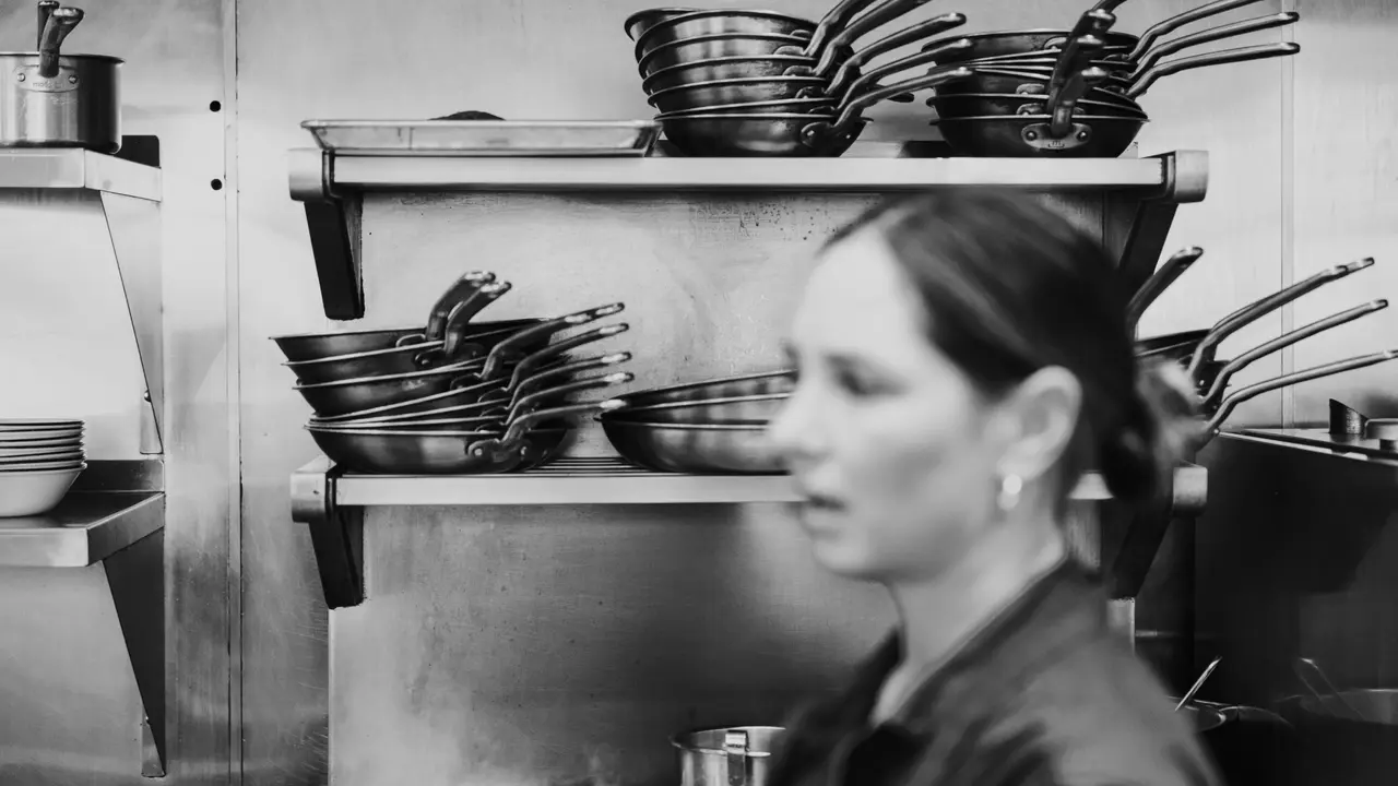 A person stands in front of shelves stocked with bowls and pans in a monochrome kitchen setting.