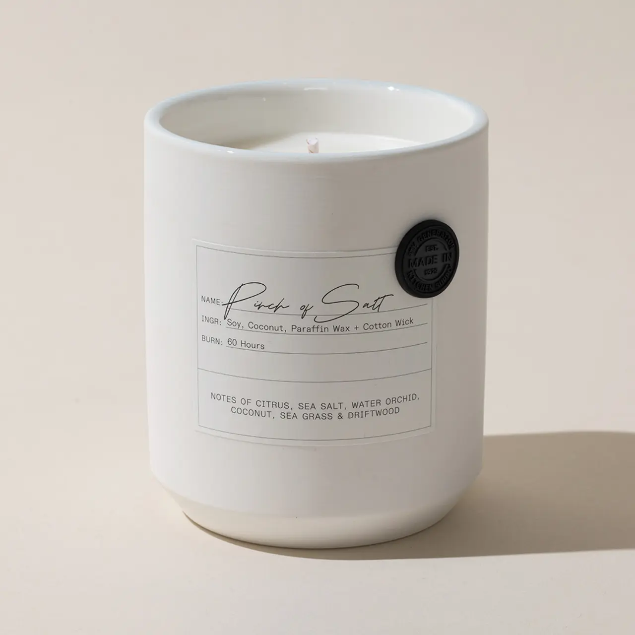 A white scented candle with a label reading "Rock & Salt," detailing ingredients and burn hours, sits against a beige background.