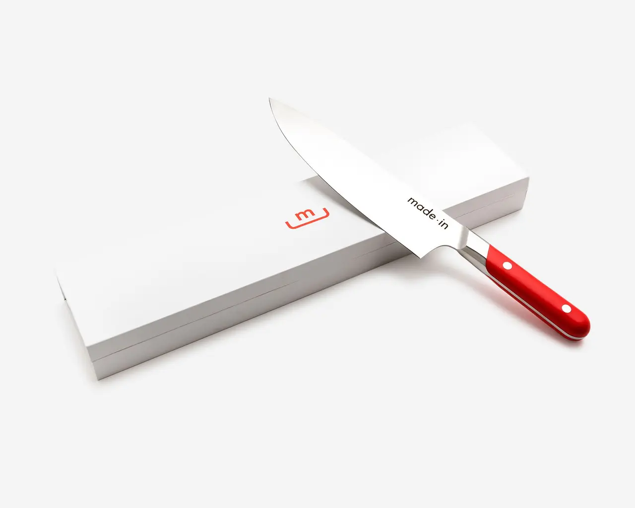 A chef's knife with a red handle lies partially out of its white packaging against a white background.