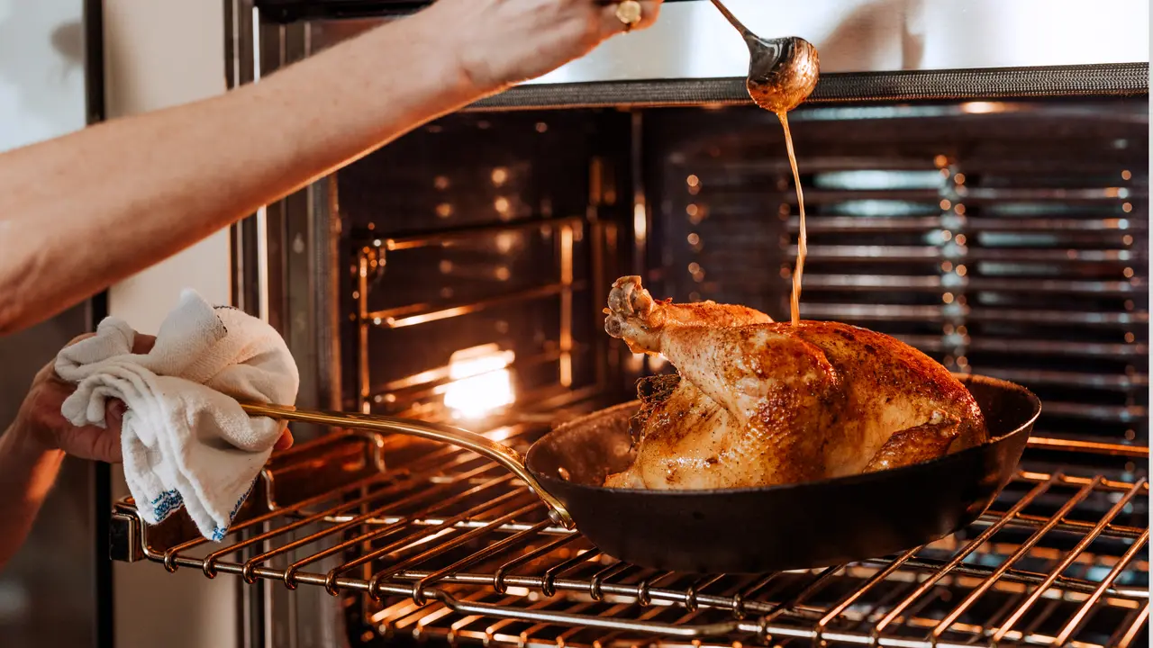 Person basting a golden-brown roasted chicken in an oven.