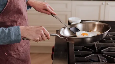 A person is cooking eggs in a skillet on a stove while wearing an apron.