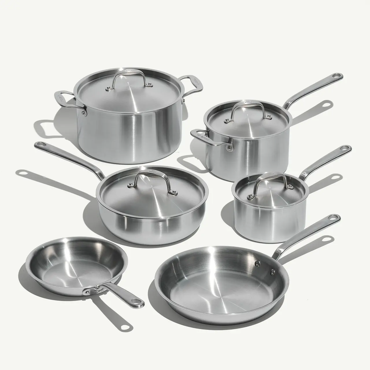 A set of stainless steel cookware including pots and pans with lids and long handles arranged on a light background.