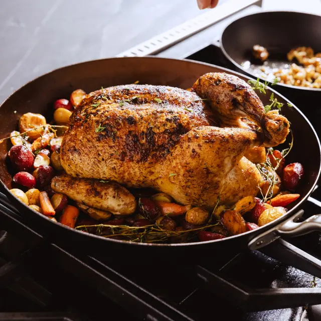 A beautifully roasted chicken is surrounded by colorful vegetables in a large skillet on a stovetop.