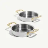 A pair of stainless steel cooking pans with gold handles sits on a light background.