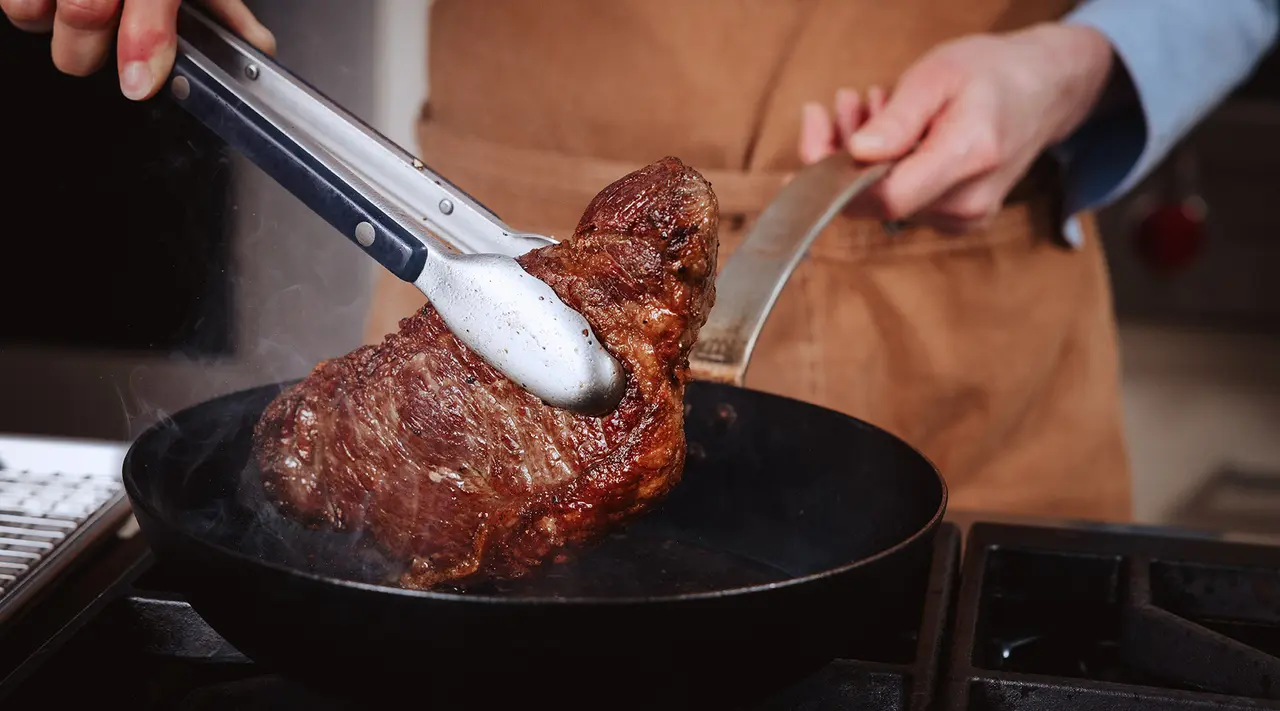 A person uses tongs to turn a large piece of meat in a sizzling skillet.