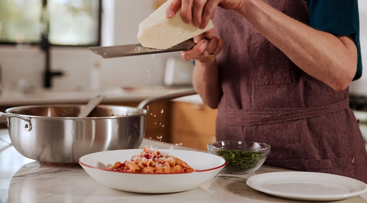 A person grates cheese over a plate of pasta in a kitchen with cooking utensils and ingredients around.