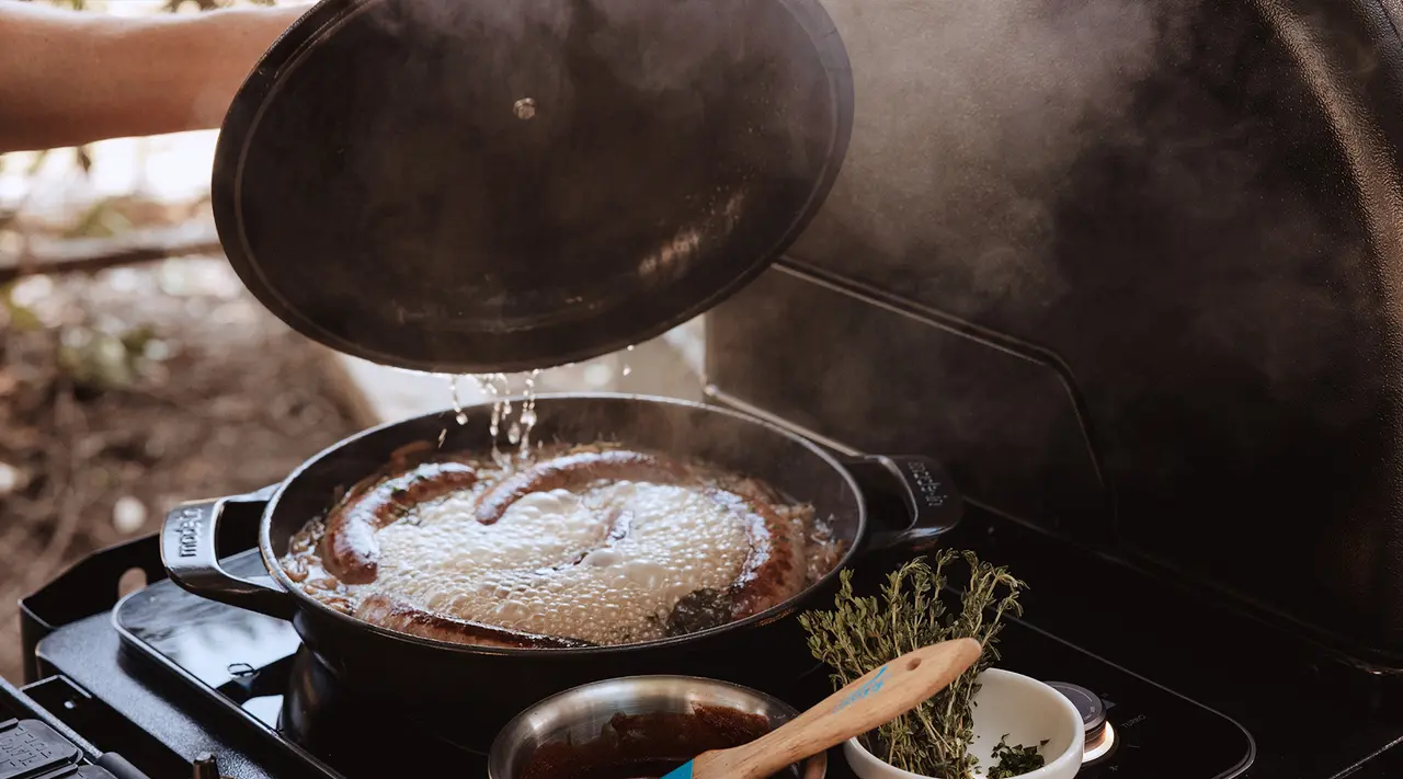 A person is cooking with a skillet outdoors, with steam rising from a bubbling sauce.