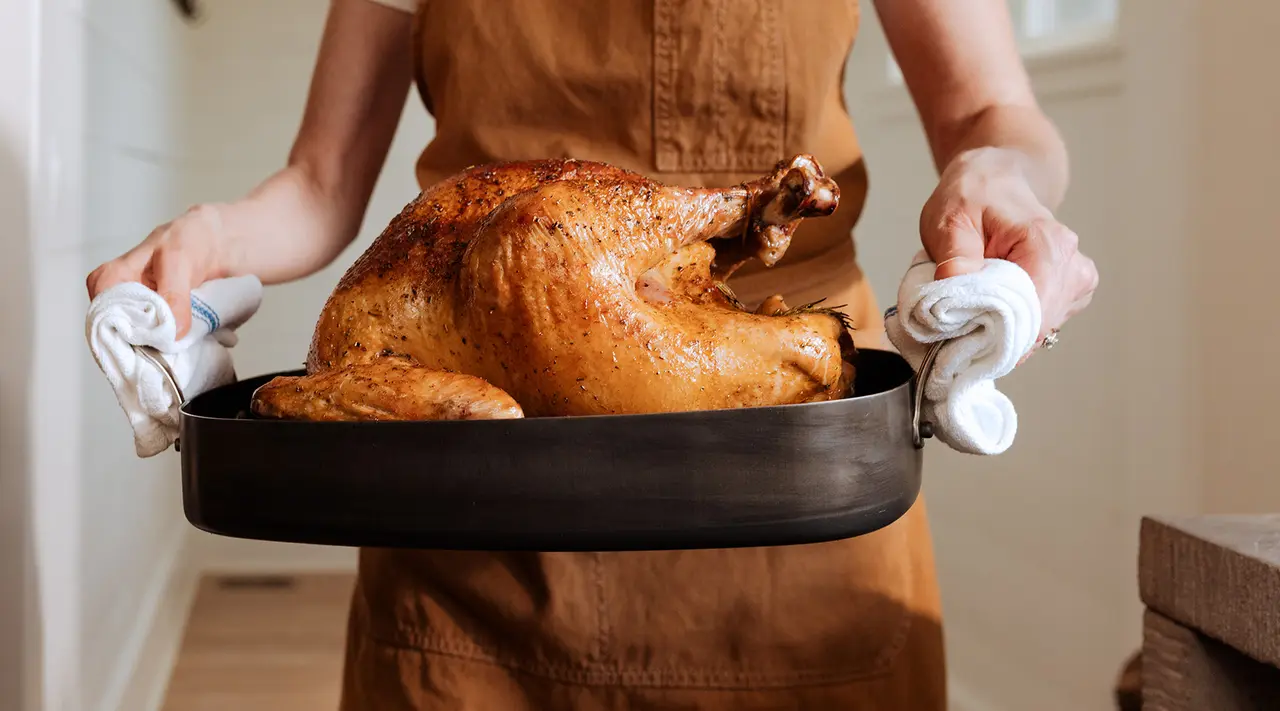 A person holding a roasted turkey in a pan with oven mitts.
