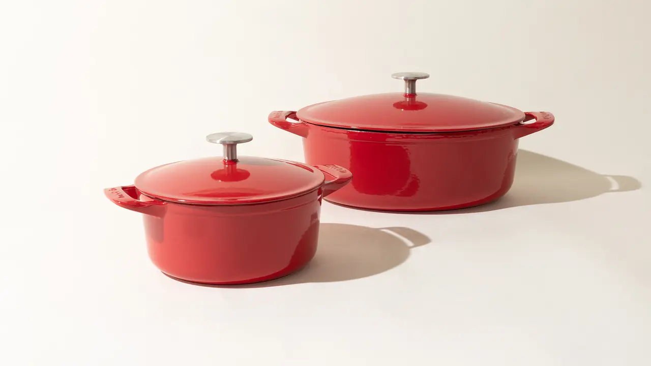 Two red enameled cast iron pots with lids, one smaller than the other, on a light background with soft shadows.