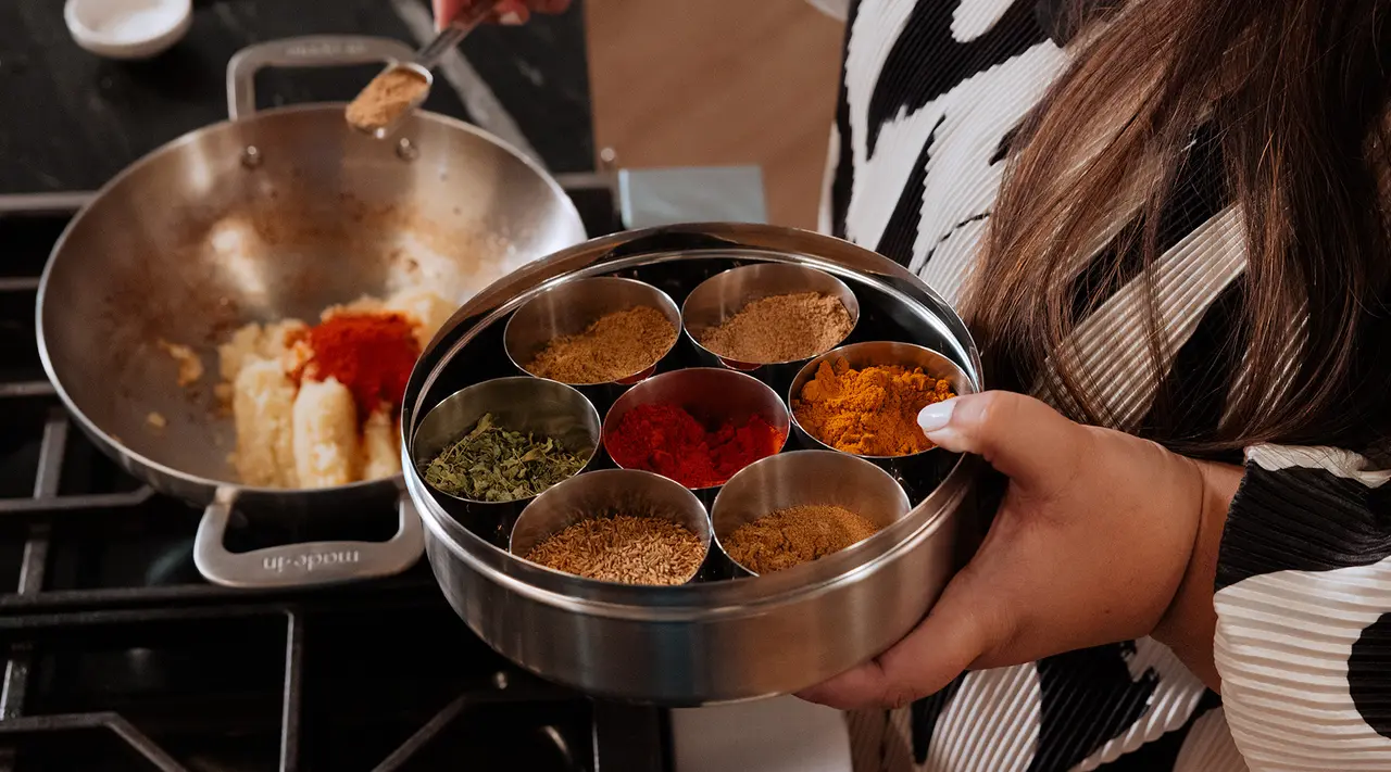 A person holds a stainless steel spice container with various colorful spices over a stove where a pan with ingredients is being cooked.