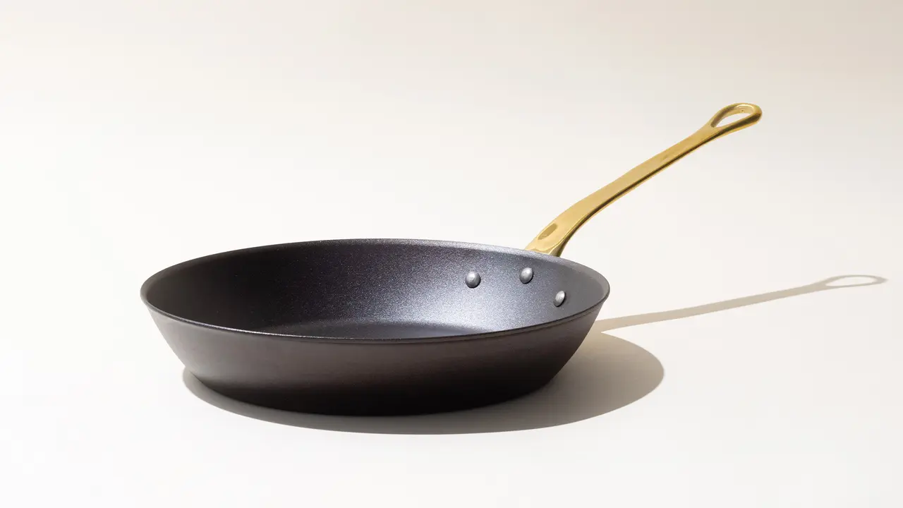 A black frying pan with a gold-colored handle on a light background, casting a shadow.