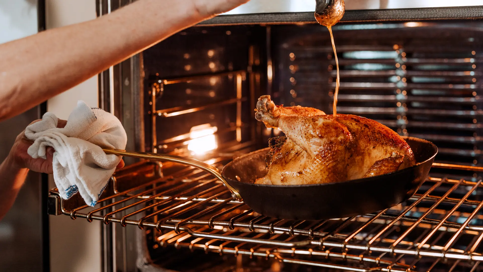 A person is basting a golden-brown roasted chicken in an oven.