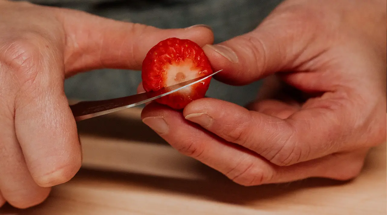 Hands are shown slicing a ripe strawberry on a wooden surface.