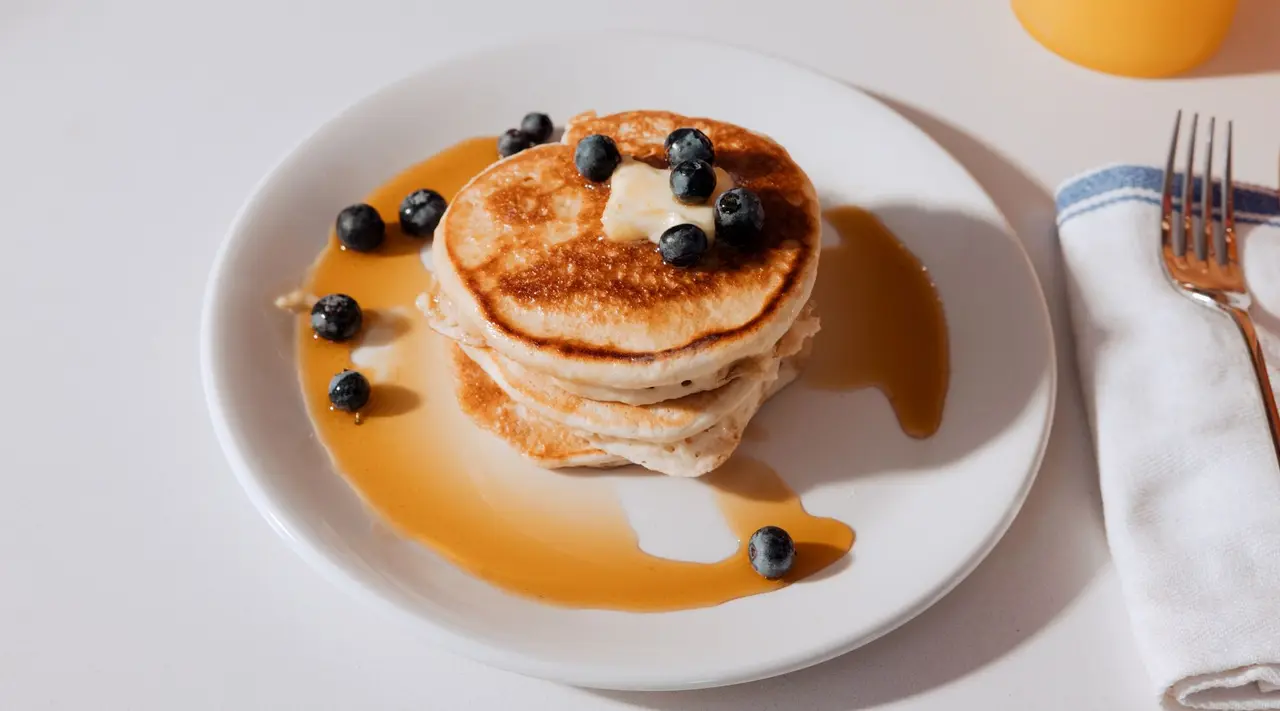 A stack of pancakes topped with blueberries and syrup is served on a plate beside cutlery and a napkin.