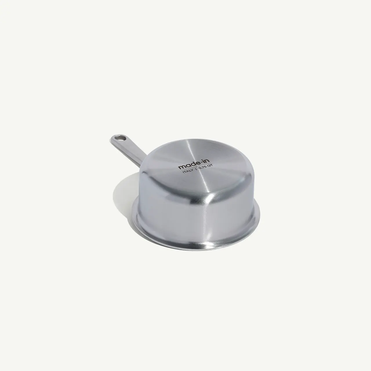 A stainless steel saucepan lid with a flat top and a looped handle sits on a matching larger plate, all against a white background.