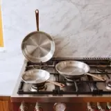 Three stainless steel frying pans, including one on the stovetop and two arranged on a marble backdrop, showcase their polished surfaces and copper handles.