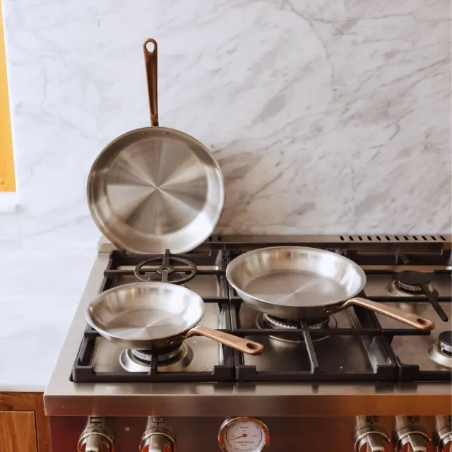 Three stainless steel frying pans, including one on the stovetop and two arranged on a marble backdrop, showcase their polished surfaces and copper handles.
