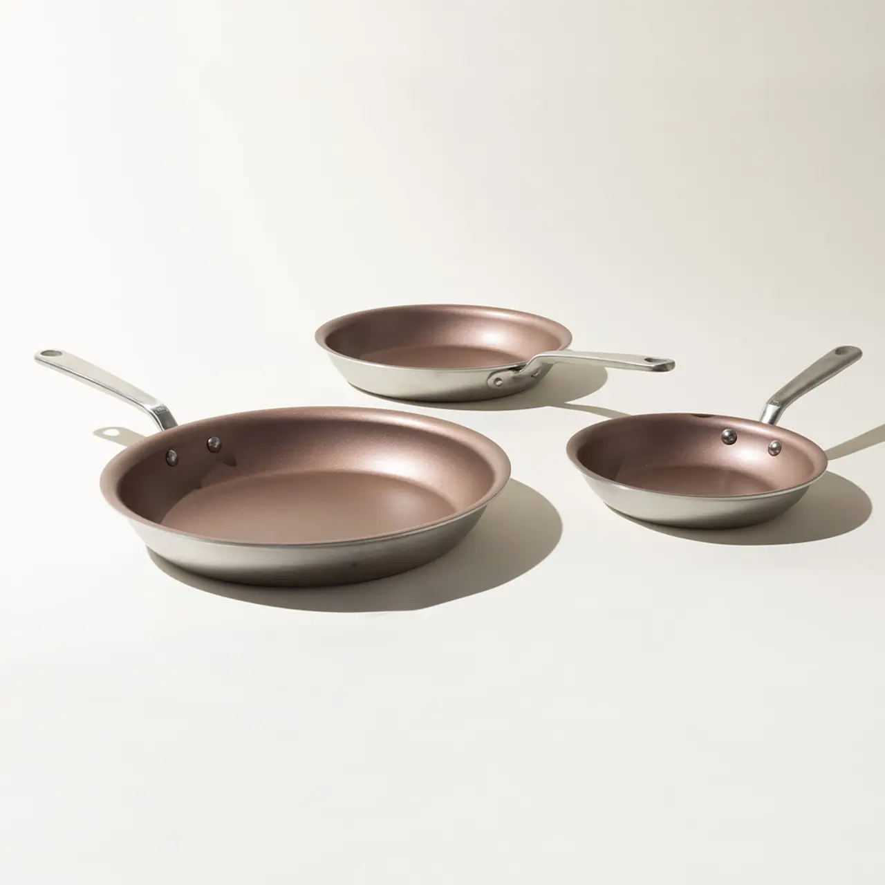 Three different-sized copper-colored frying pans with silver handles are arranged on a light background.