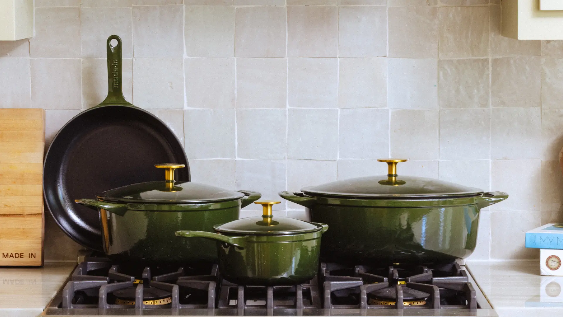 Three green enameled cast iron pots and a skillet are arranged on a stovetop against a light-tiled backsplash.