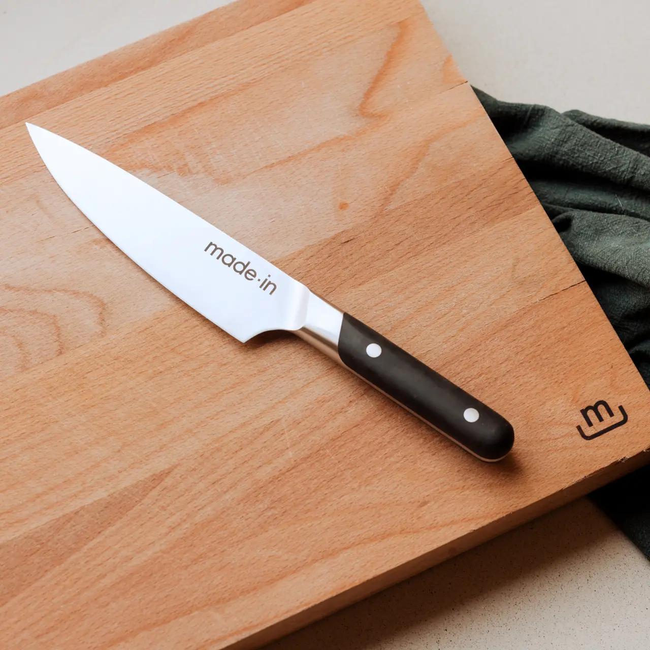 A chef's knife with a black handle lies on a wooden cutting board next to a dark green cloth.