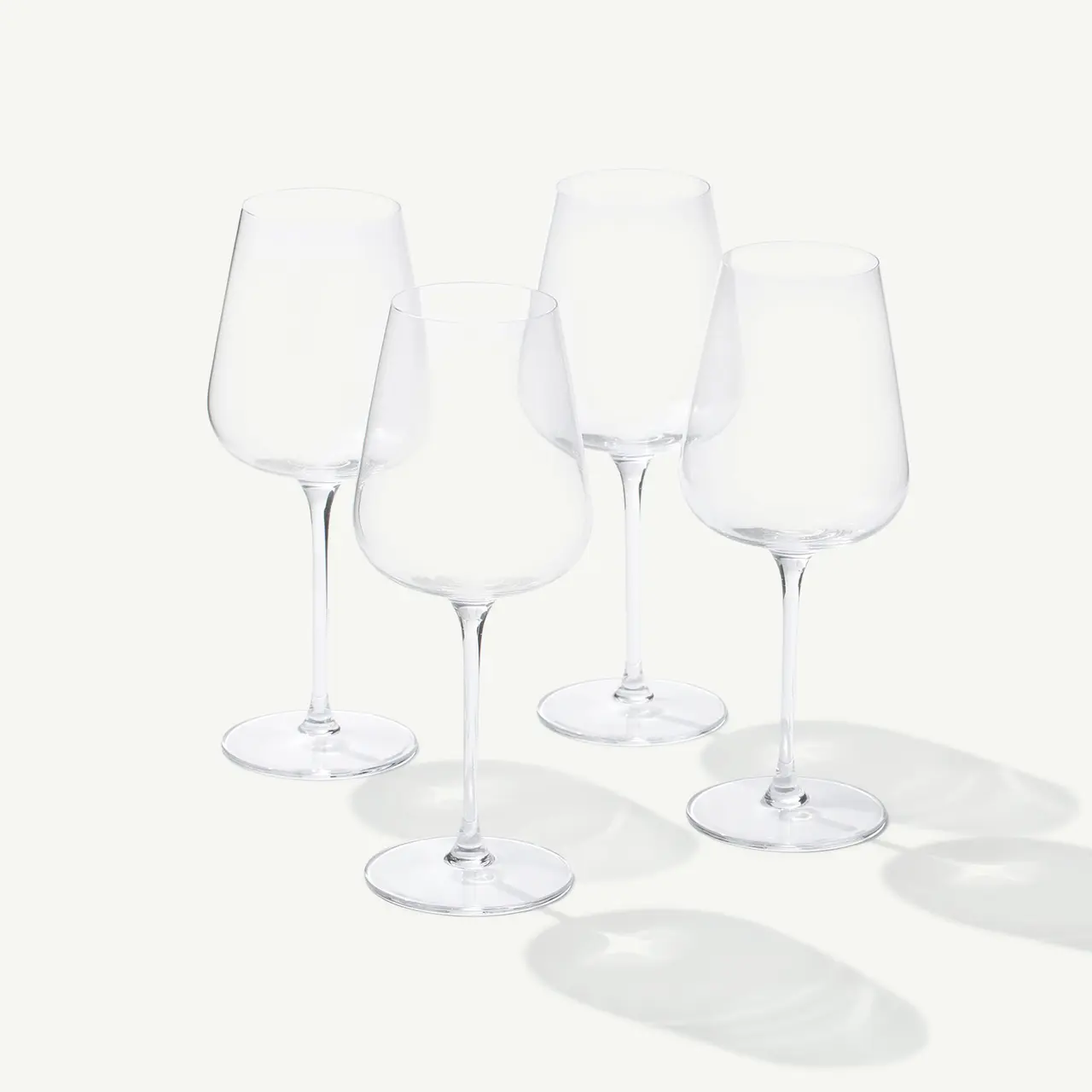 Four empty wine glasses are positioned upright on a white surface with shadows visible beneath them.