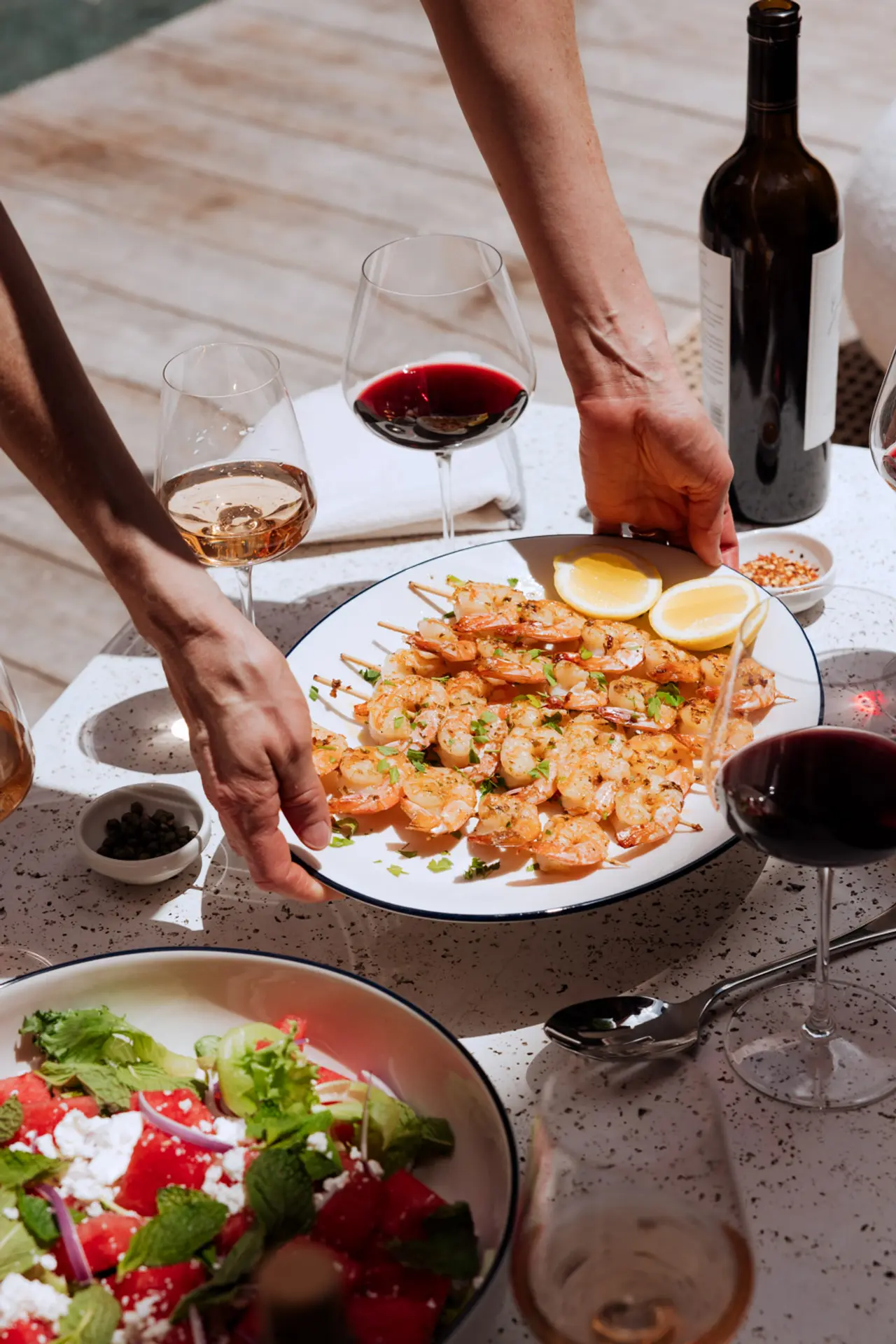 Hands serve shrimp on a plate amidst a table with wine glasses, a bottle, and a bowl of salad, evoking a sense of a social dining experience.