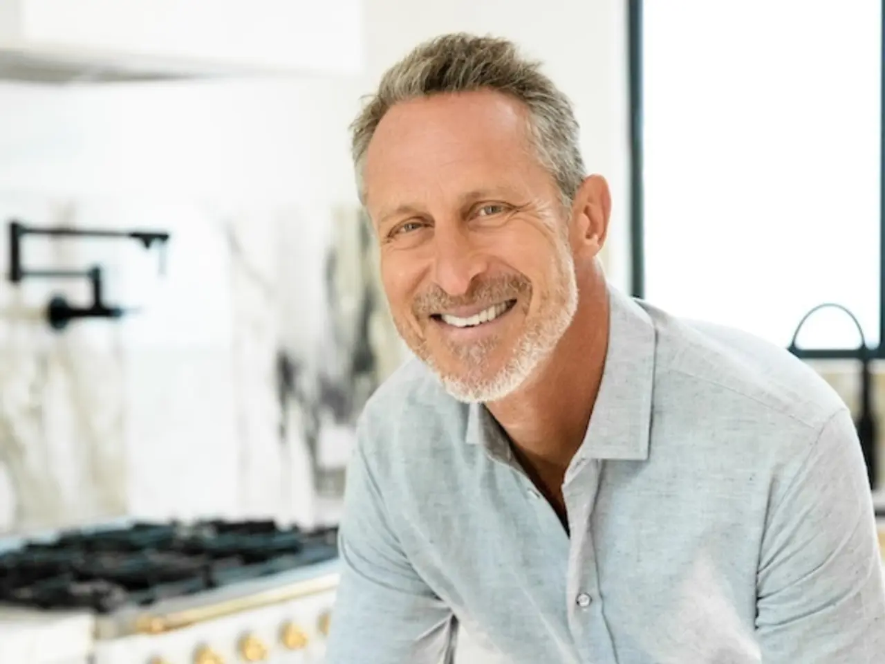 A smiling man with short gray hair and a beard is posed in a modern kitchen setting.