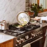 A person is sautéing vegetables and lemon slices in a stainless steel frying pan on a gas stove in a modern kitchen.