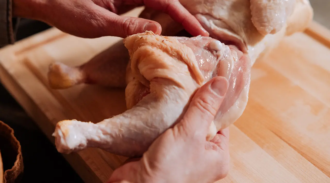 Hands are preparing a raw chicken on a wooden cutting board for cooking.