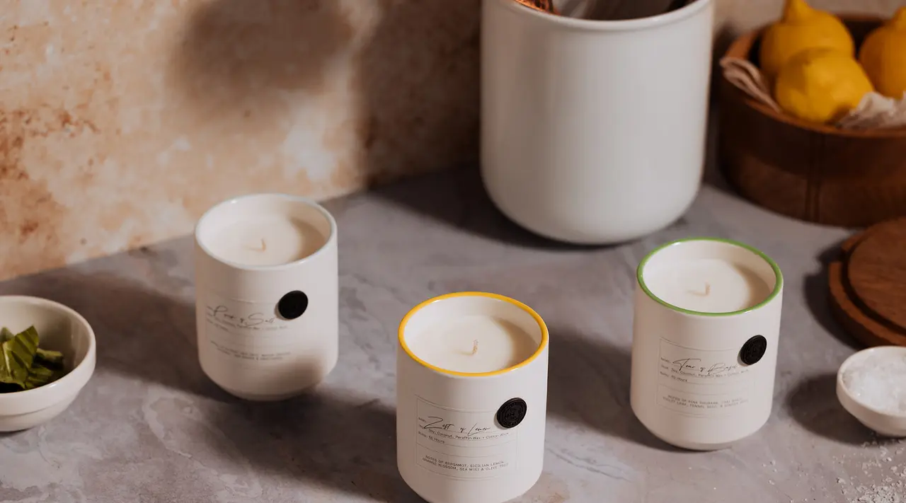Scented candles with stylish labels are displayed on a textured surface alongside kitchen bowls and utensils.