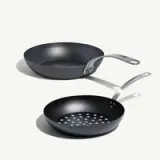 A black non-stick frying pan and a slotted frying pan with stainless steel handles are displayed on a light background.