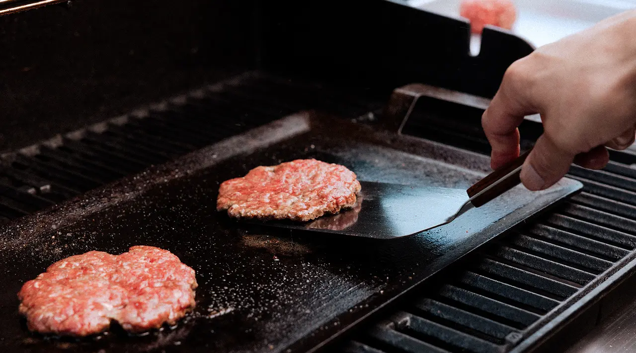 A person's hand flipping burgers on a hot grill with a spatula.