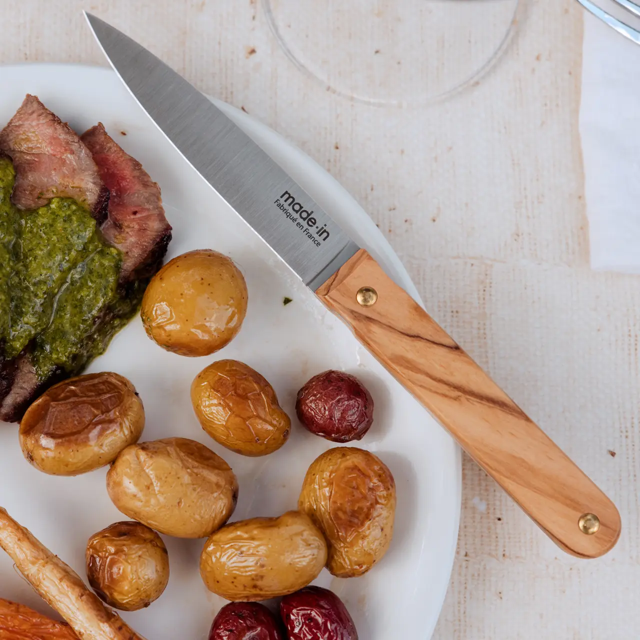A chef's knife rests next to a plate of roasted potatoes and sliced steak with a dollop of green sauce.