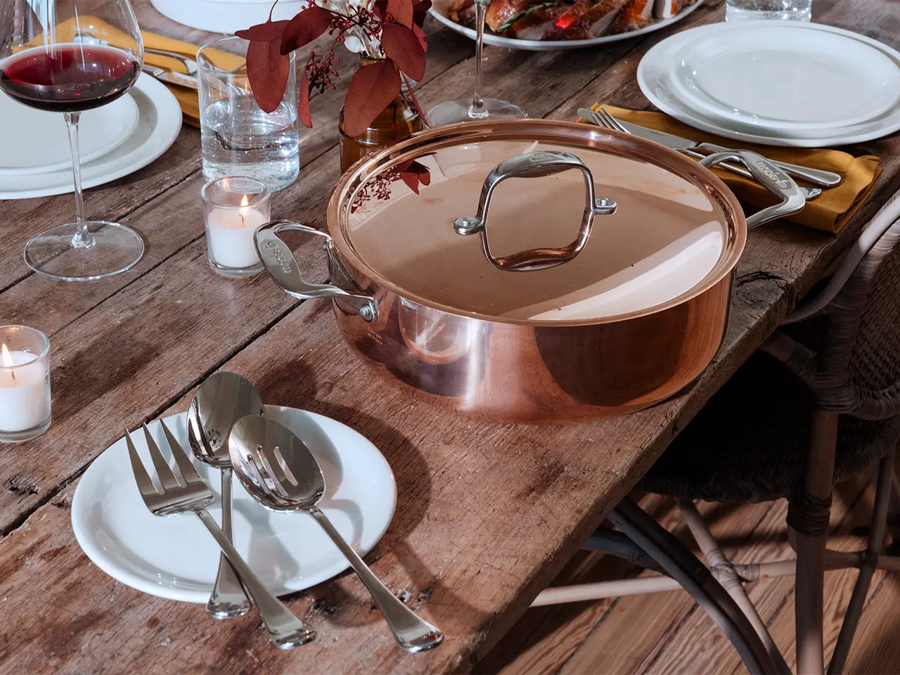A copper pot sits on a dining table set with plates, cutlery, and glasses of wine, ready for a meal.