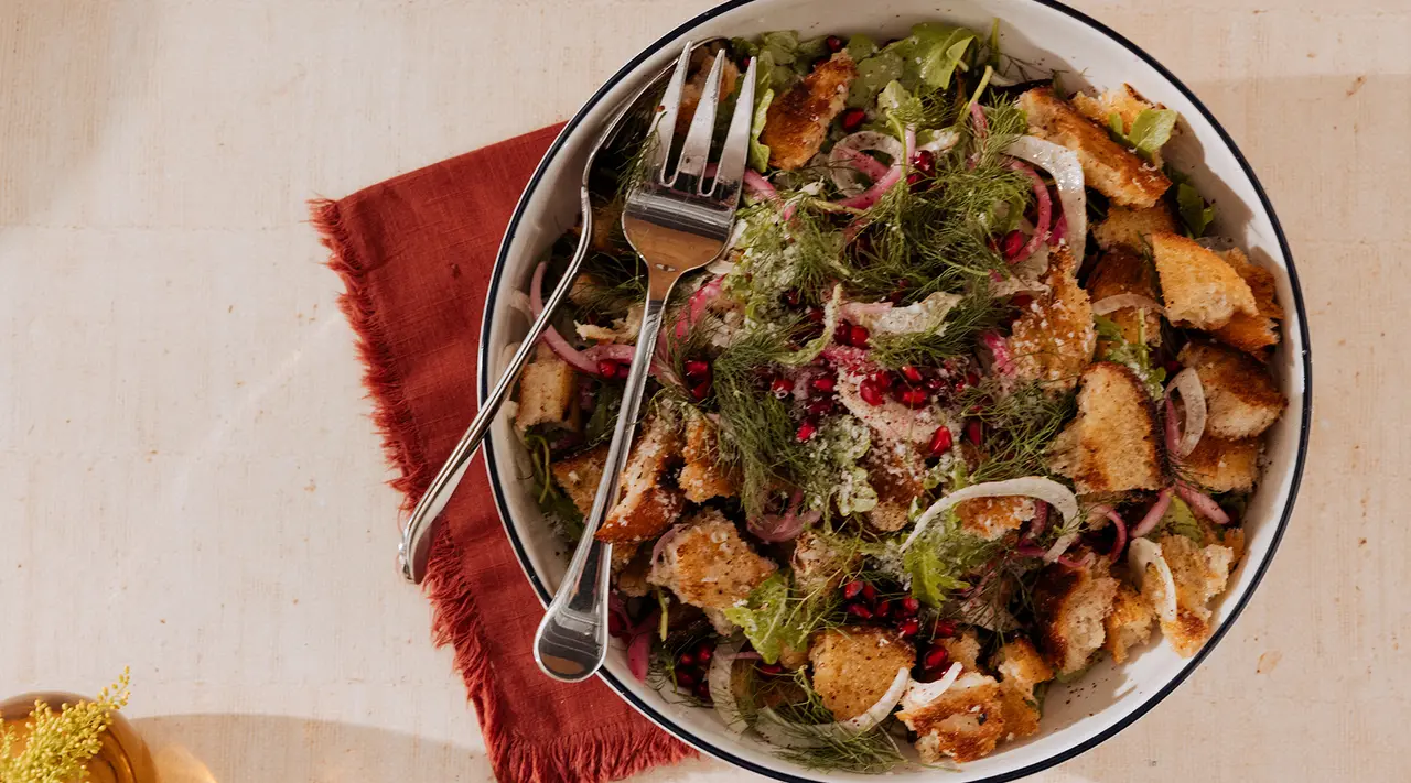 A bowl of panzanella, an Italian bread salad, is presented with chunks of bread, red onion, leafy greens, and a sprinkling of herbs and pomegranate seeds.