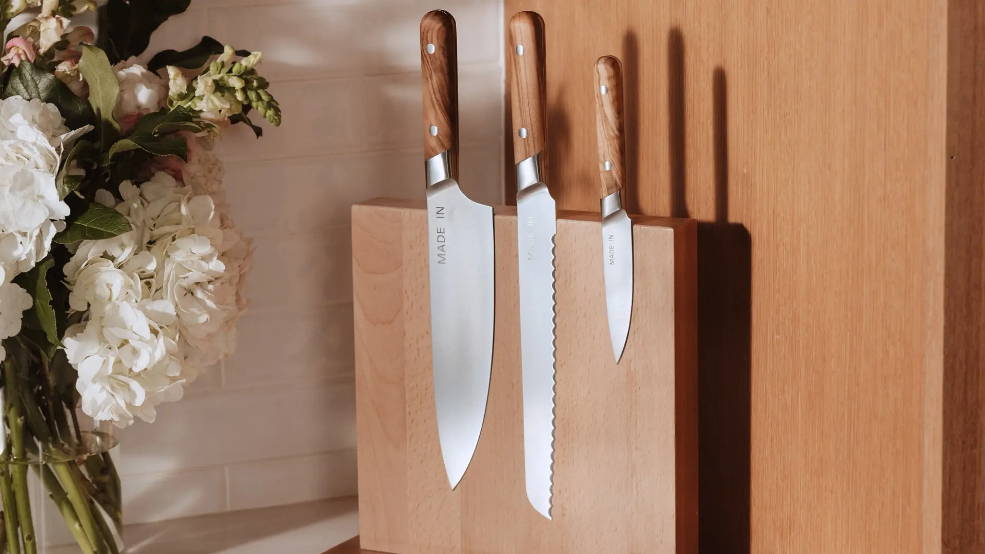 Three kitchen knives with wooden handles are displayed in a wooden knife block alongside a bouquet of flowers.