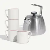 Four white mugs with red rims stacked beside a stainless steel kettle.