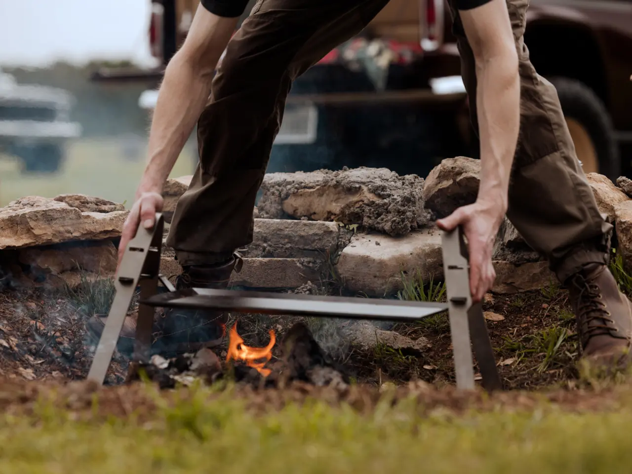 A person in brown pants and boots is stoking a small fire with a long-handled tool outdoors, near a stone ring and a vehicle.